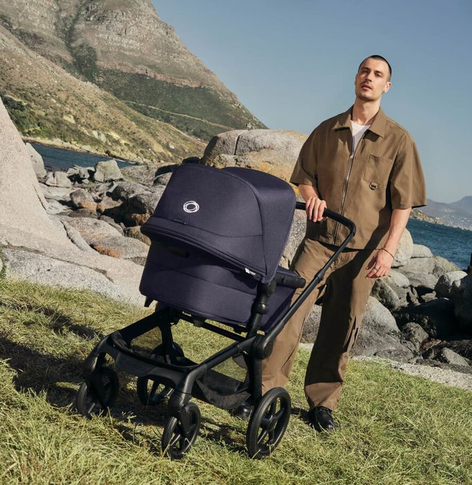 A dad in a casual brown outfit pushes a Bugaboo Fox 5 Renew newborn stroller on grassy terrain. Behind him are large rocks and a scenic coastal backdrop.