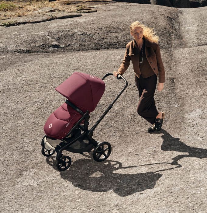 A fashionably dressed mom walks with a Bugaboo Fox 5 Renew seat stroller in Dark Cherry colorway on rocky terrain. The stroller has a footmuff in the same color.