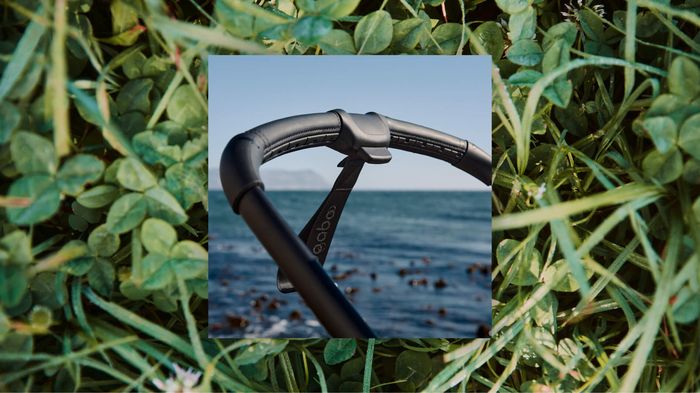 An image of the handlebar on the Bugaboo Fox 5 Renew stroller with a view of the ocean behind it. It's superimposed on a second image, which is a close-up of green leaves on the ground.