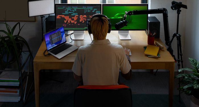 Person sitting at desk with a mic and headphones in front of three devices, Mac Studio Display screen, audio editing tools, sound clips in different blocks of color, a second Mac Studio Disp