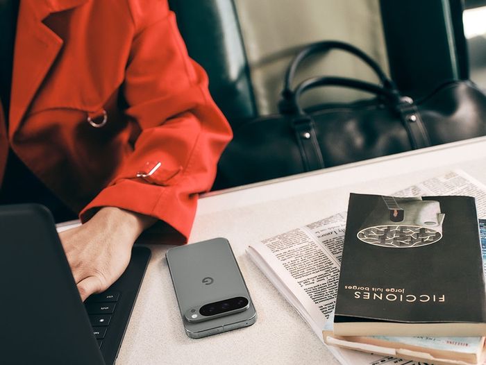 Google Fi Wireless - An individual sits at a desk with a laptop, a newspaper and a book. A Pixel 9 in Moonstone is faced down on the desk.
