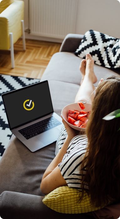 Woman lying on a couch eating a bowl of fruit with a laptop beside her displaying Norton logo